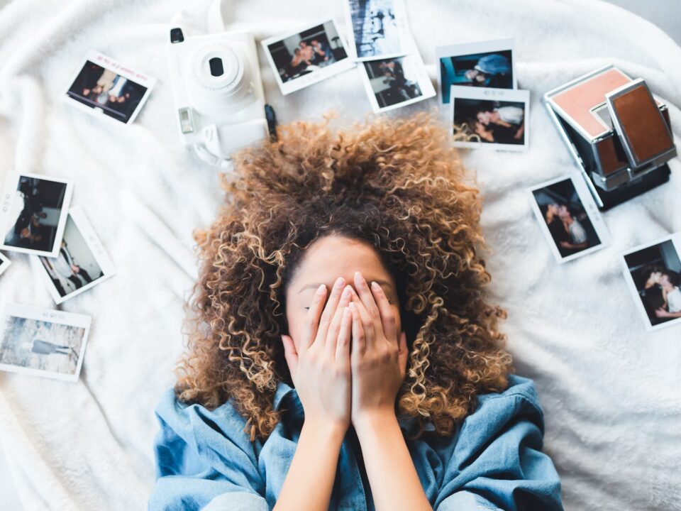 woman lying on bed covering her face surrounded by photos and white camera