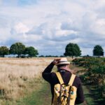 man holding fedora cap while walking on green and beige grass field under white clouds