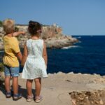 two children standing near cliff watching on ocean at daytime