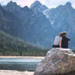 a couple sits on a rock looking out over a lake