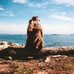 woman in brown sweater sitting on rock near sea during daytime