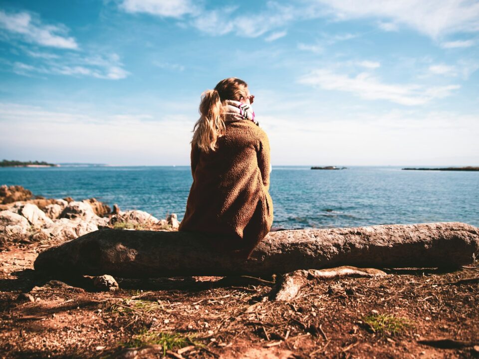 woman in brown sweater sitting on rock near sea during daytime
